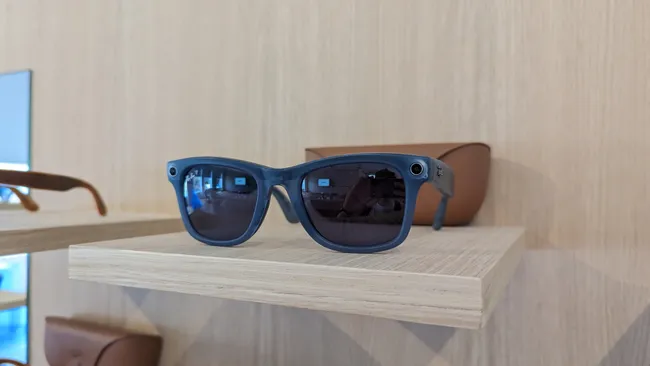 Tendencias en Gafas Inteligentes con IA | XionGadget 5 Meta Rayban con marco Azul sobre soporte de madera y de fondo su estuche cargador sobre pared de madera